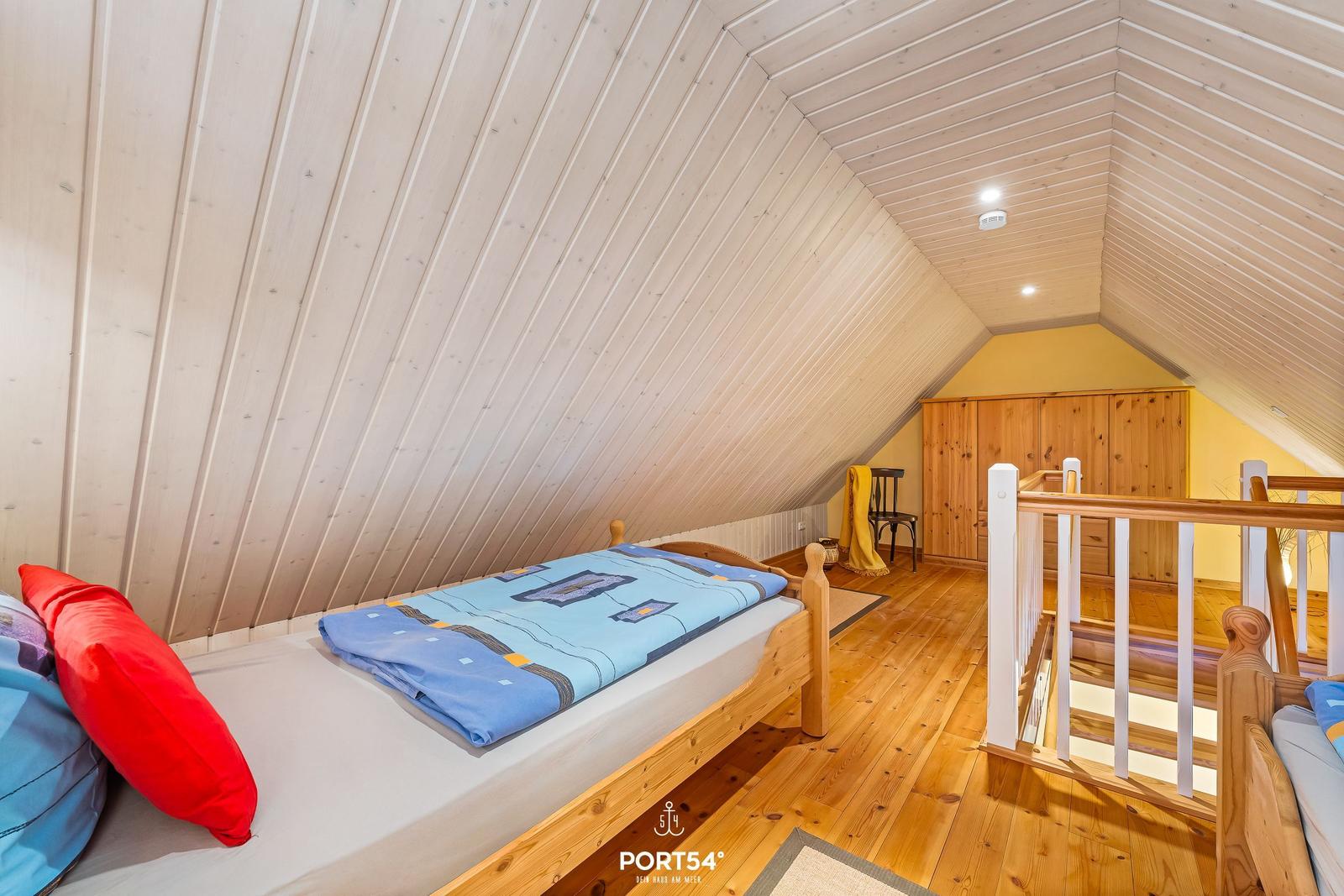 Schlafzimmer mit Holzboden und Dachschrägen. Bett mit blauer Bettwäsche und rotem Kissen.