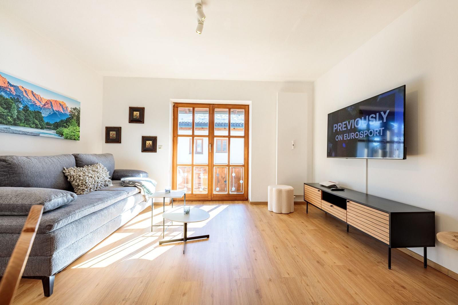 Wohnzimmer mit Sofa, TV und Holzboden. Fenster zur Terrasse.