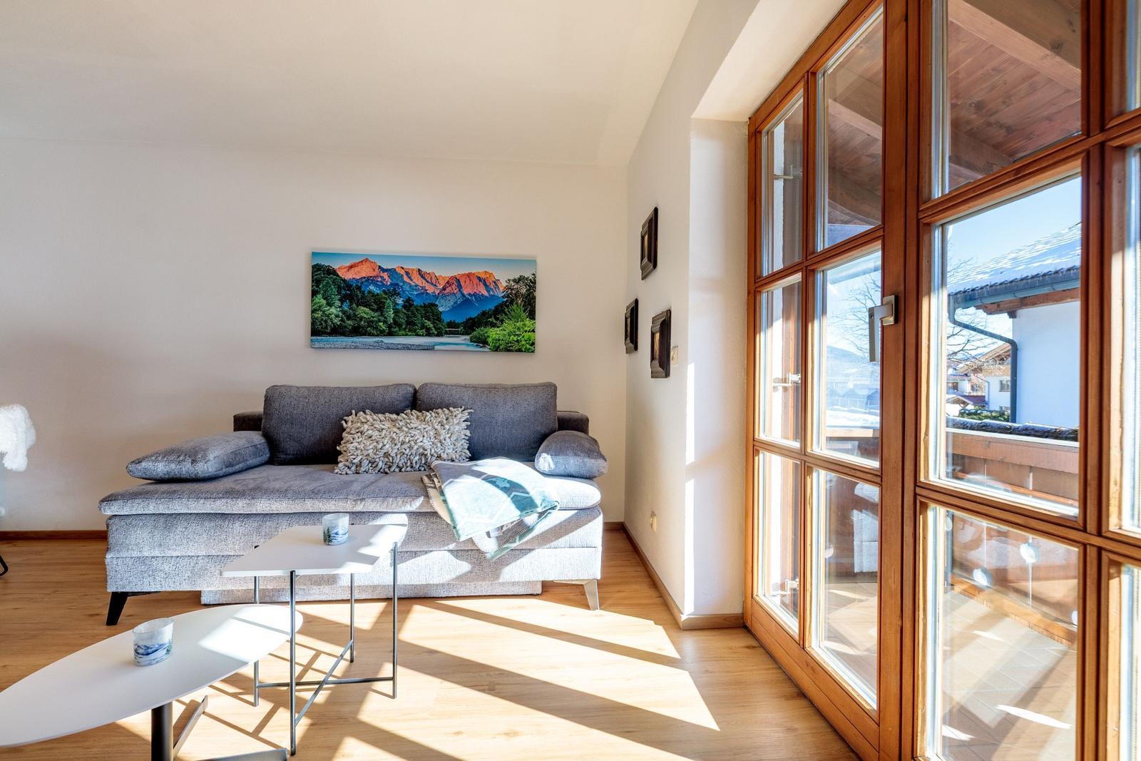 Wohnzimmer mit Sofa, Tischen und großem Fenster. Wandbild zeigt Berge.
