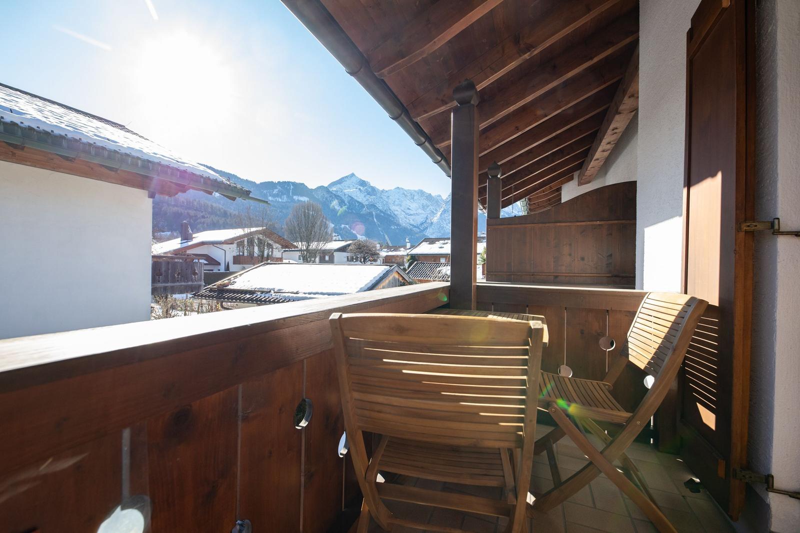 Terrasse mit Holzgeländer und zwei Stühlen. Blick auf verschneite Berge und Dächer.