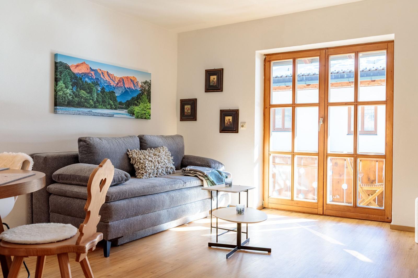 Wohnzimmer mit Sofa, Tischen und Holzboden. Große Glastür führt auf Balkon.