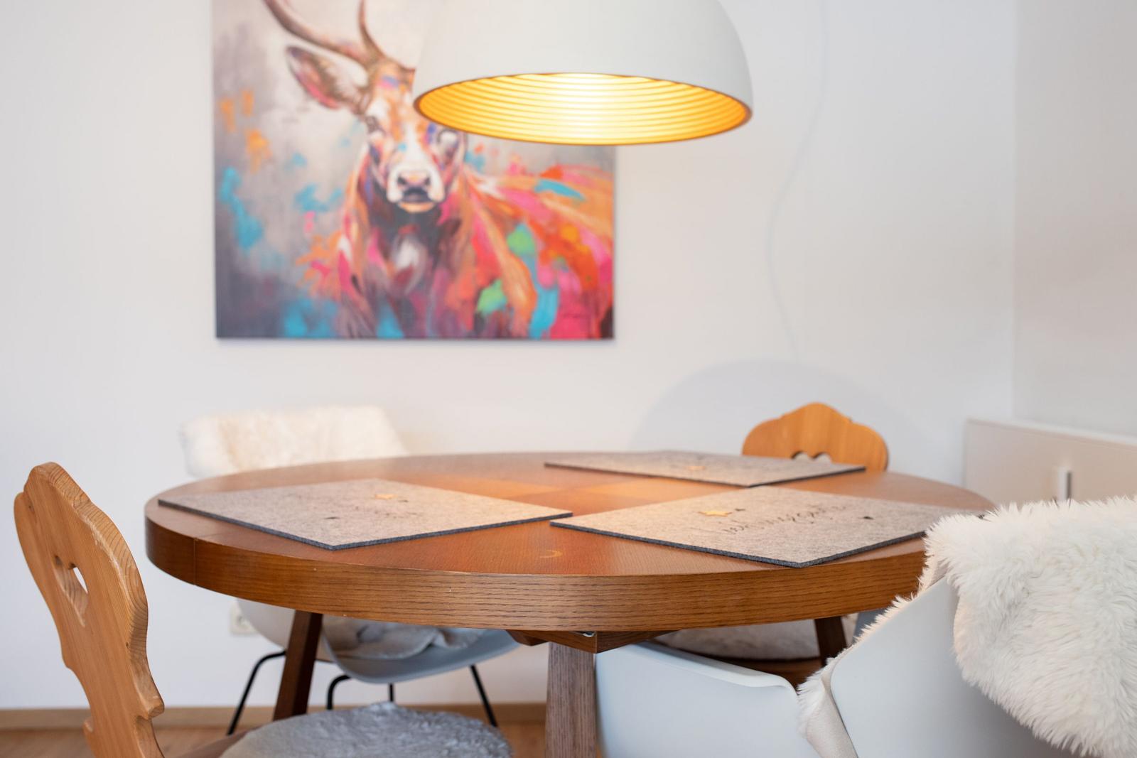 Runder Holztisch mit Stühlen und Platten unter einer Lampe. Wandbild zeigt ein farbenfrohes Reh.