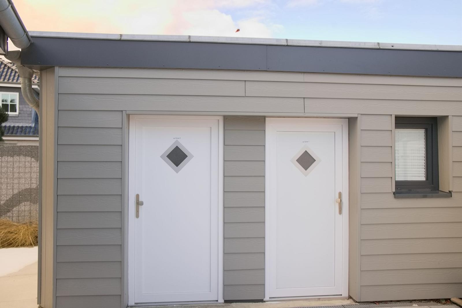 Zwei weiße Türen mit Diamantfenstern an einem Holzhaus mit grauer Fassade.
