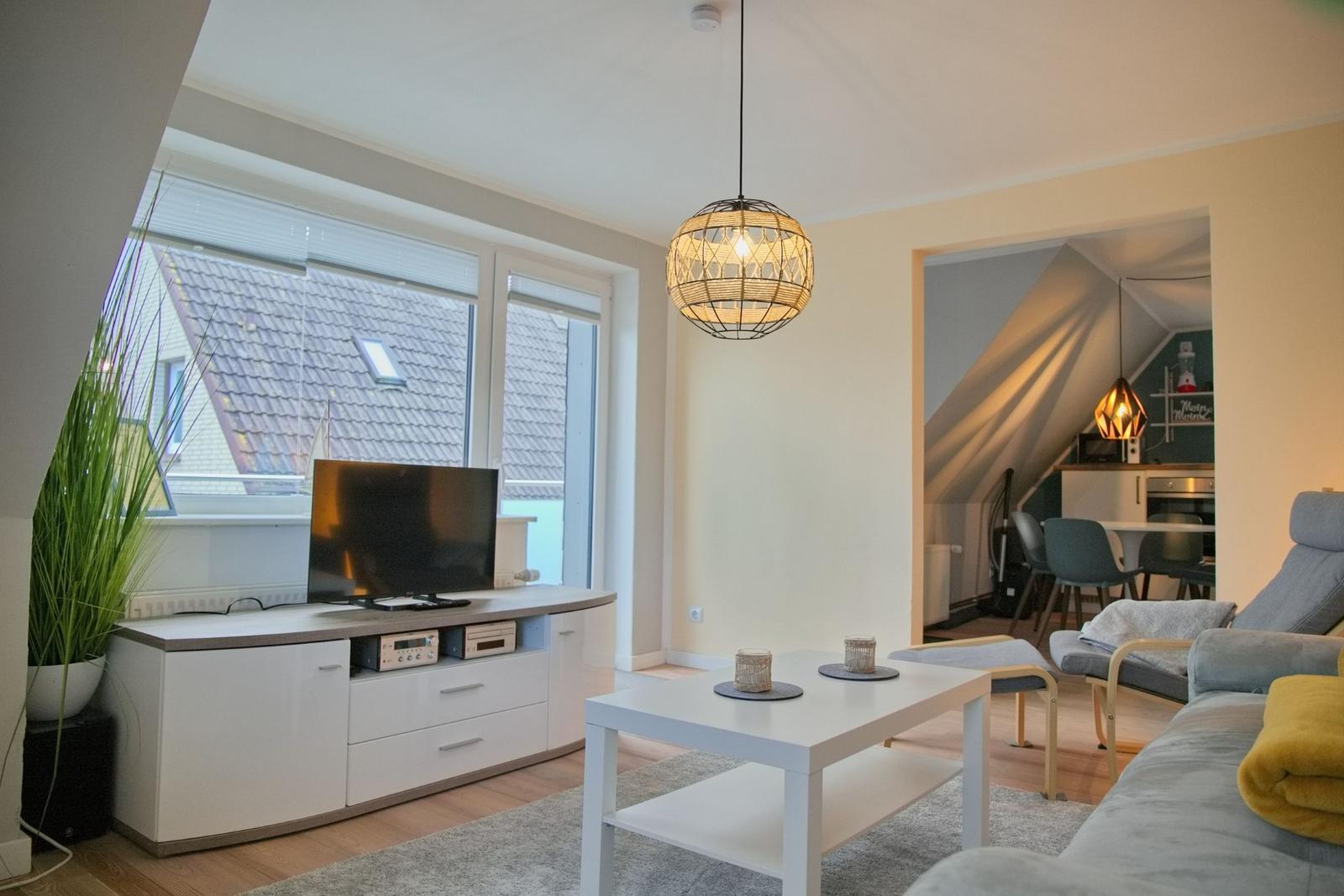 Wohnzimmer mit TV, Couch und Tisch. Küche im Hintergrund sichtbar.