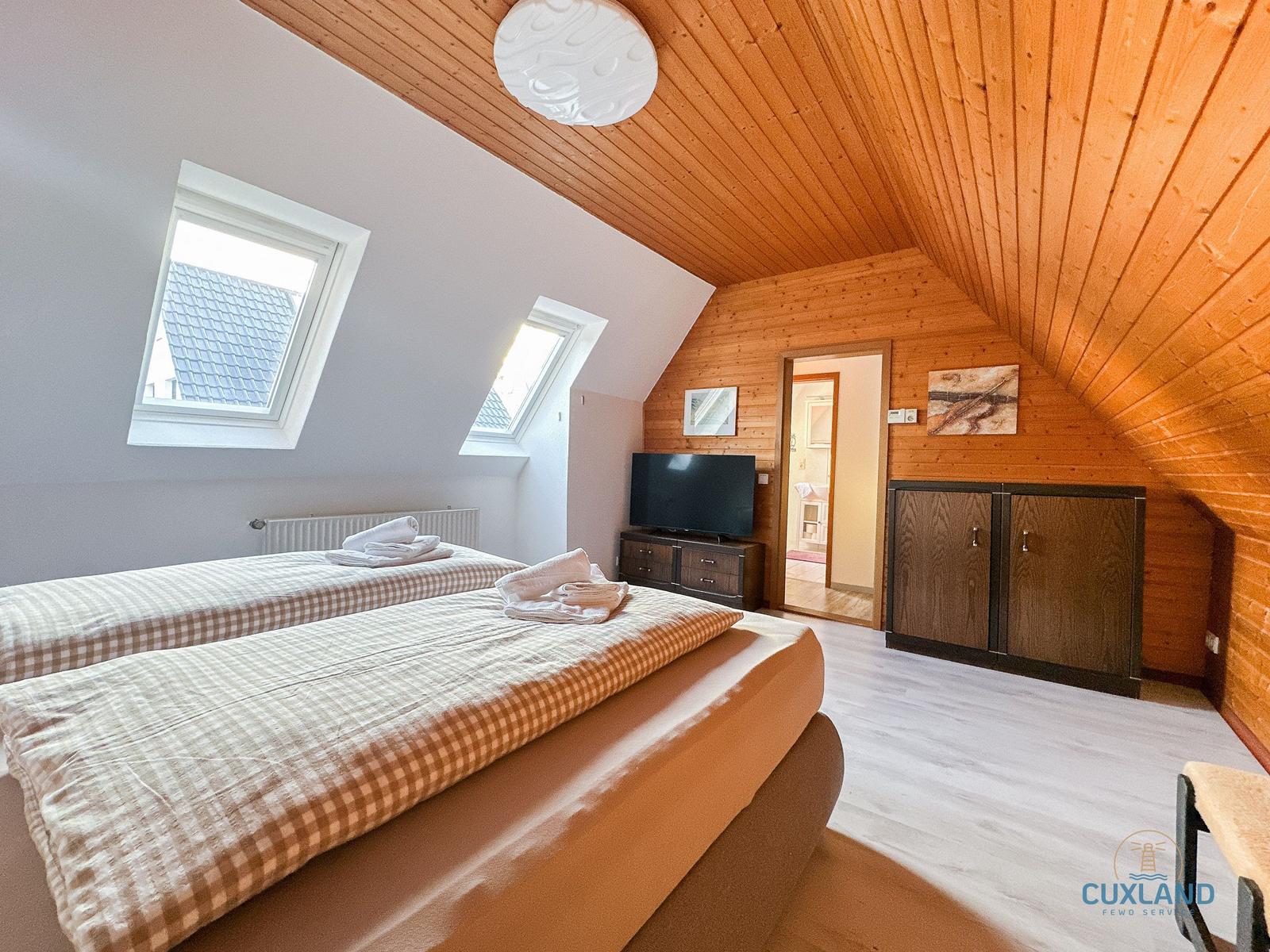 Schlafzimmer mit Dachschrägen, zwei Fenstern und Bett mit Tisch.