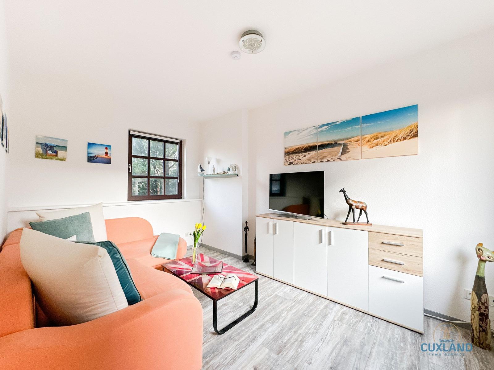 Ein Wohnzimmer mit orangefarbenem Sofa, TV-Schrank und Strandmotiven an der Wand.