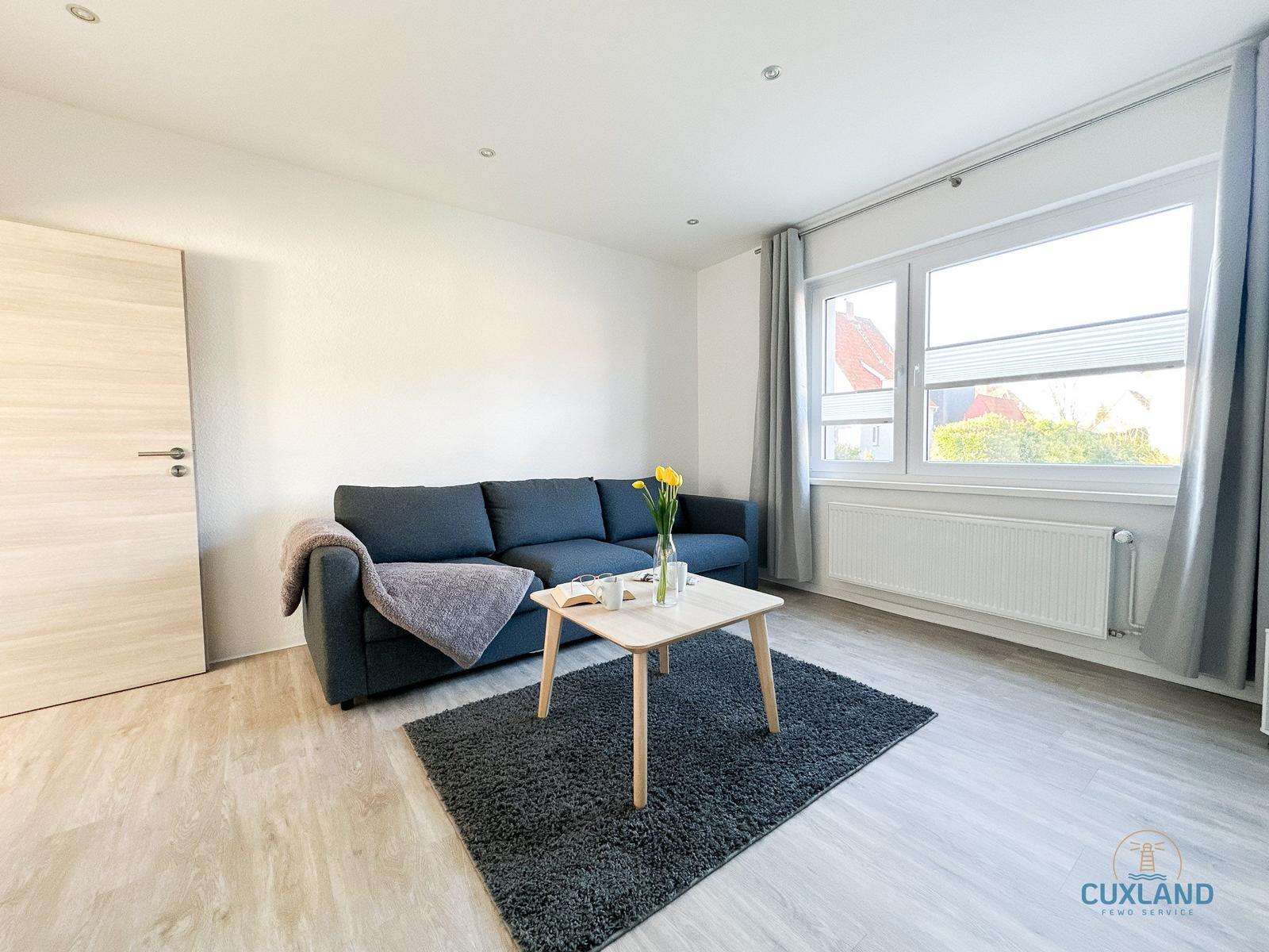 Wohnzimmer mit Sofa, Tisch und Fenster. Holzboden und graue Gardinen.