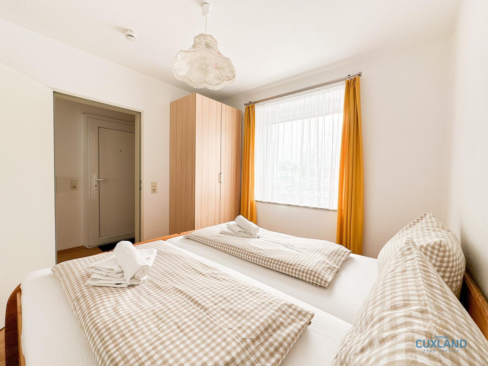 Zwei Betten mit karierten Bettwäschen, Fenster mit gelben Vorhängen und Holzschrank.