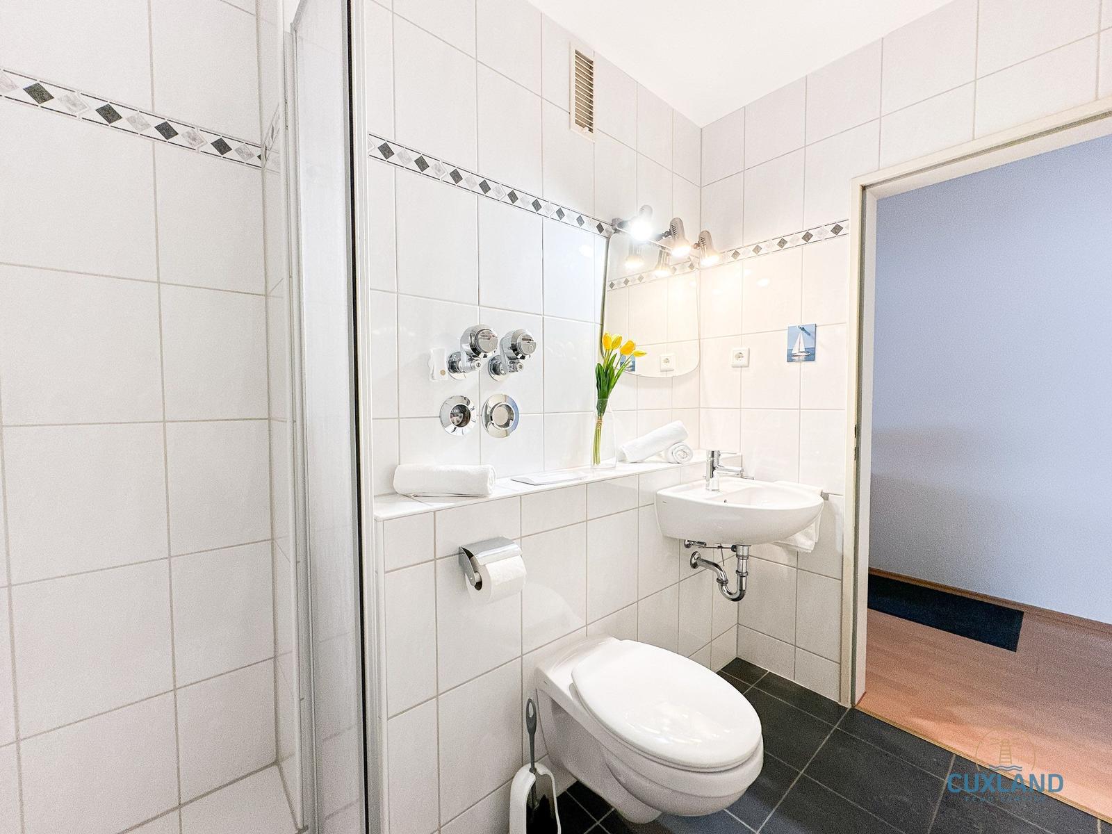 Badezimmer mit Dusche, Waschbecken und Toilette. Weiße Fliesen und schwarze Bodenfliesen.