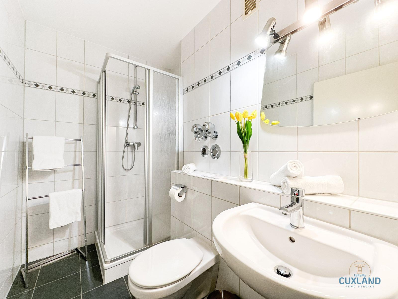 Badezimmer mit Dusche, Waschbecken und Toilette. Weiße Fliesen und gelbe Tulpen.