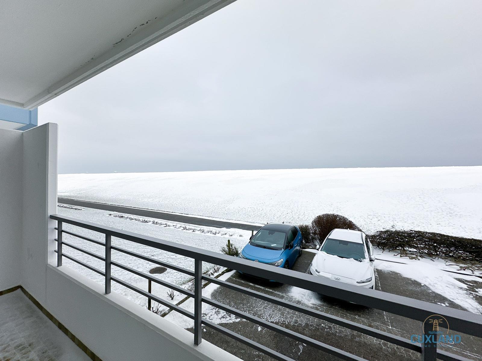 Blick vom Balkon auf schneebedecktes Gelände mit zwei parkenden Autos.