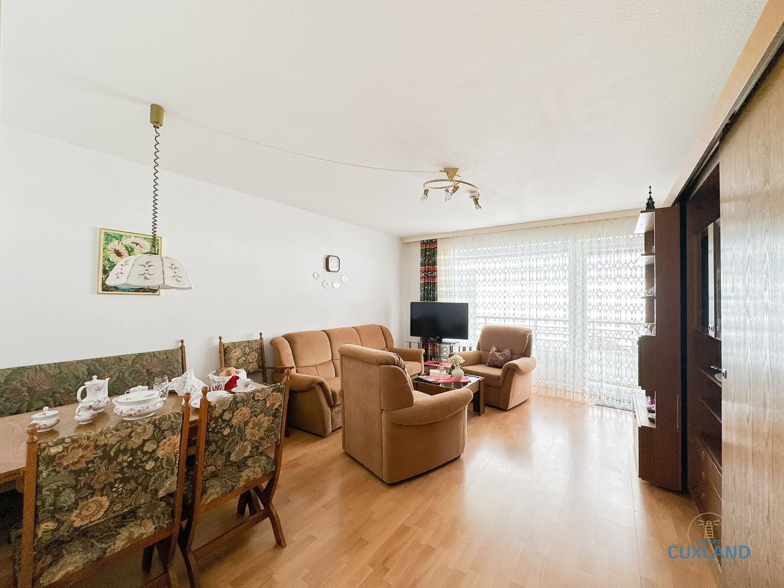 Wohn- und Essbereich mit Sofa, Sesseln und Esstisch. Holzboden und Fenster mit Vorhängen.