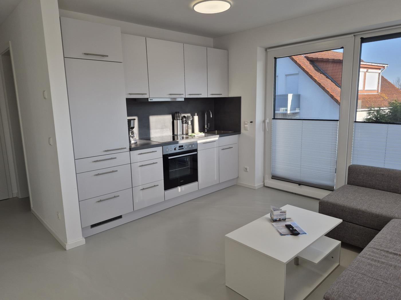 Küche mit weißen Schränken und integriertem Ofen. Wohnbereich mit grauem Sofa und Fenster.