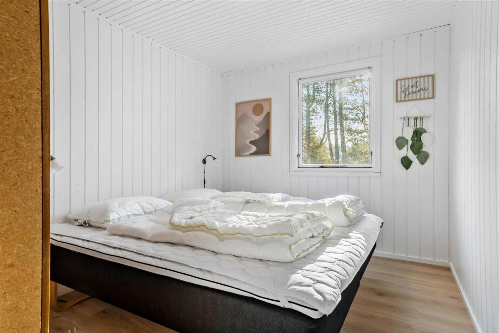 Schlafzimmer mit weißem Bett, Fensterblick auf Wald und Wanddekoration.