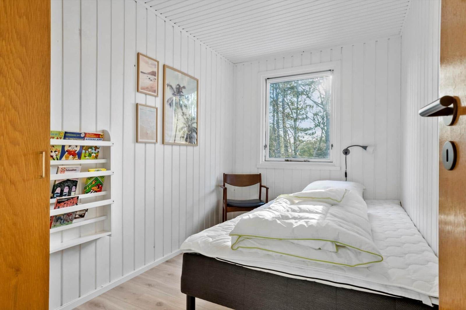 Schlafzimmer mit Bett, Stuhl, Fenster und Bücherregal an der Wand.