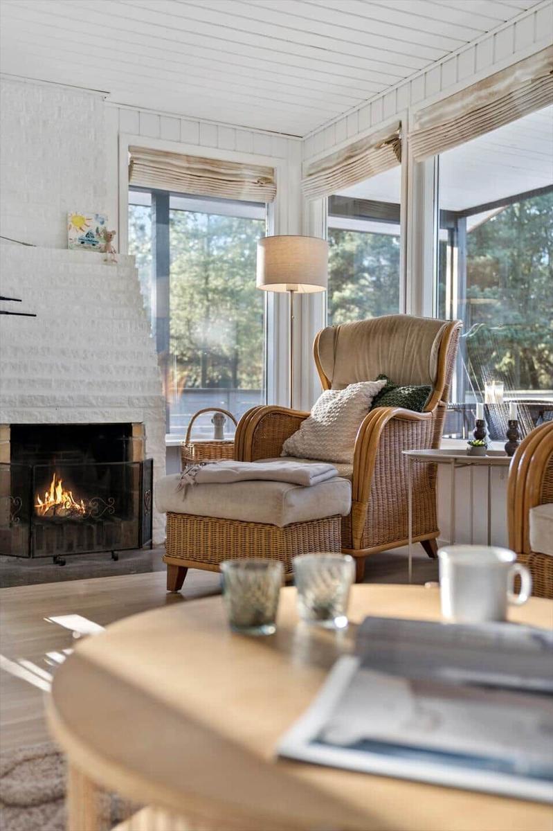 Wohnzimmer mit Kamin, Sessel und Tisch. Große Fenster mit Blick auf Wald.