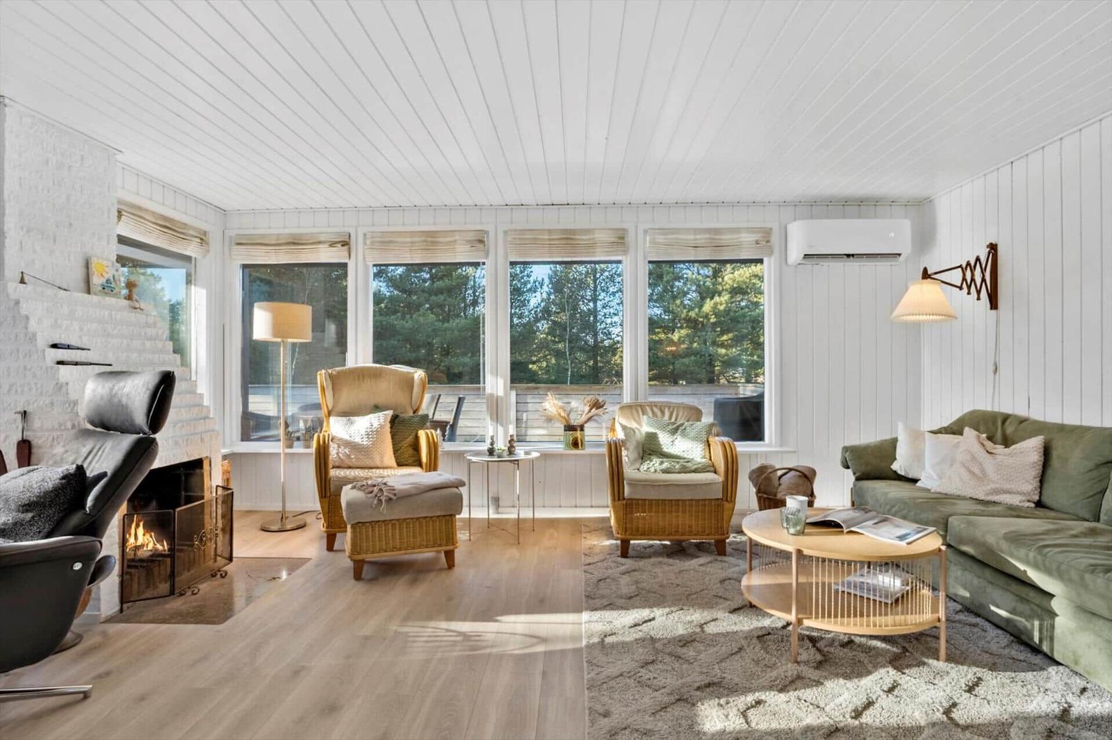 Wohnzimmer mit Kamin, Sofa, Sesseln und großen Fenstern mit Blick auf Wald.