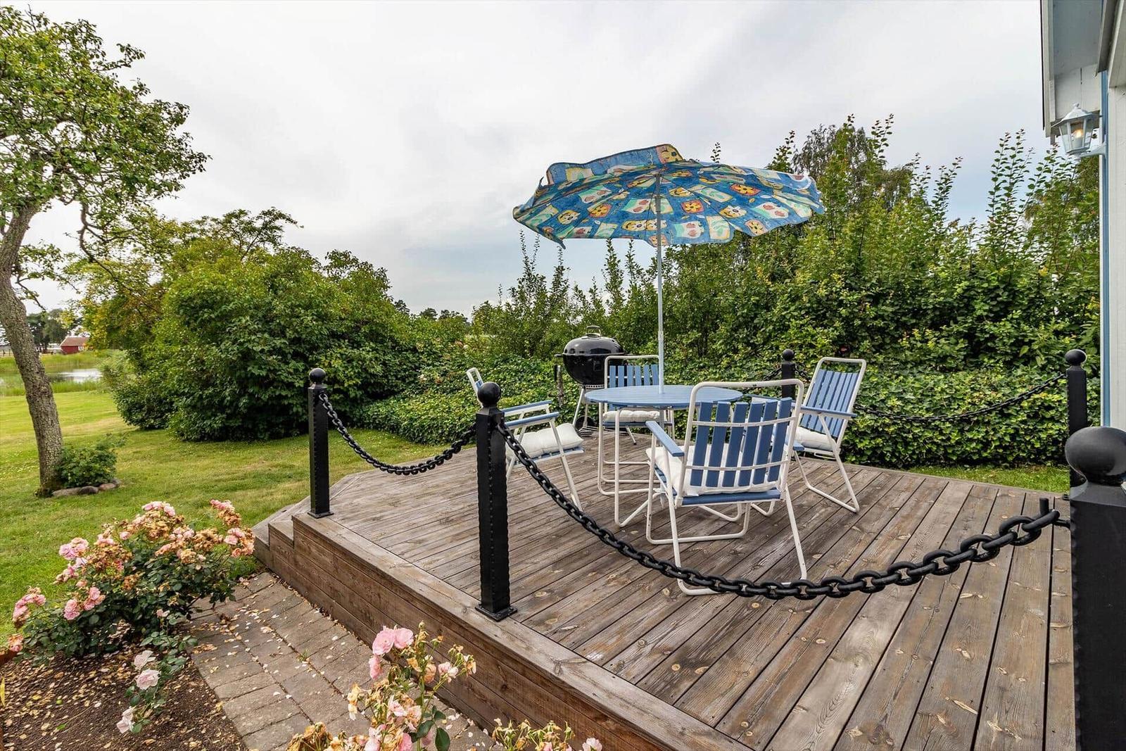 Terrasse mit Tisch, Stühlen, Grill und Sonnenschirm. Umgeben von Grün und Blumen.