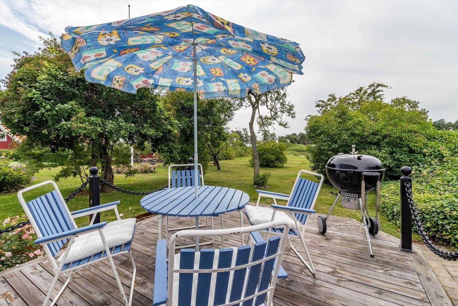 Terrasse mit Tisch, Stühlen, Sonnenschirm und Grill. Umgeben von Grün und Bäumen.