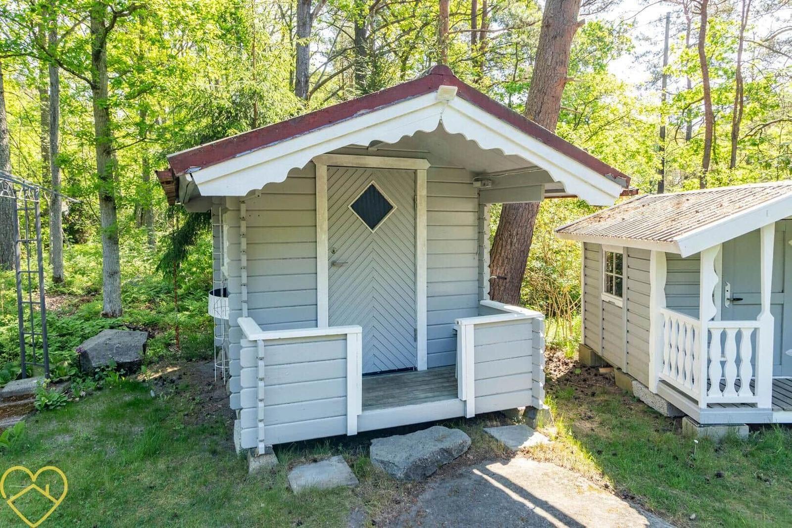 Zwei kleine Holzhütten mit Veranda im Wald. Eine mit Dachgaube, die andere mit Schiebetüren.