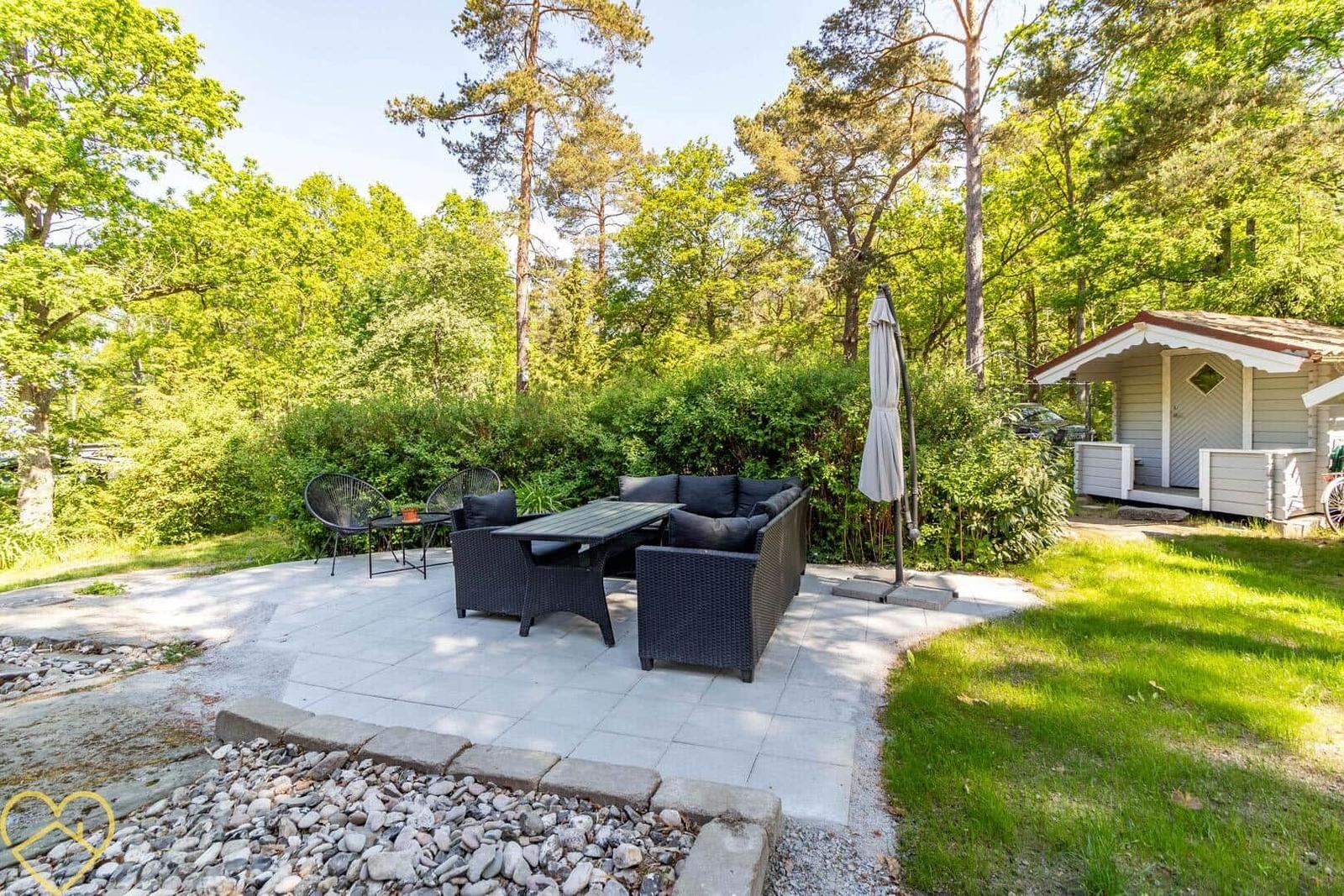 Terrasse mit Sitzgruppe, Tisch und Sonnenschirm. Hintergrund: Wald und Gartenhaus.