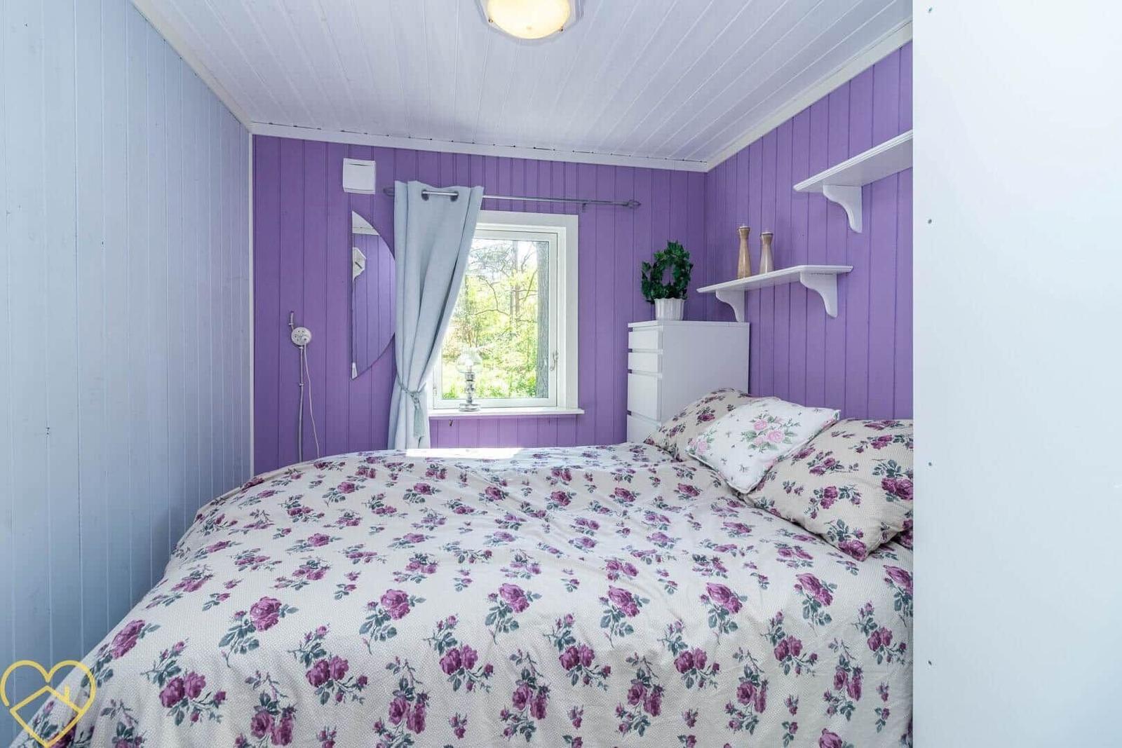 Schlafzimmer mit lila verkleideten Wänden, Fenster und Bett mit Blumenbezug.