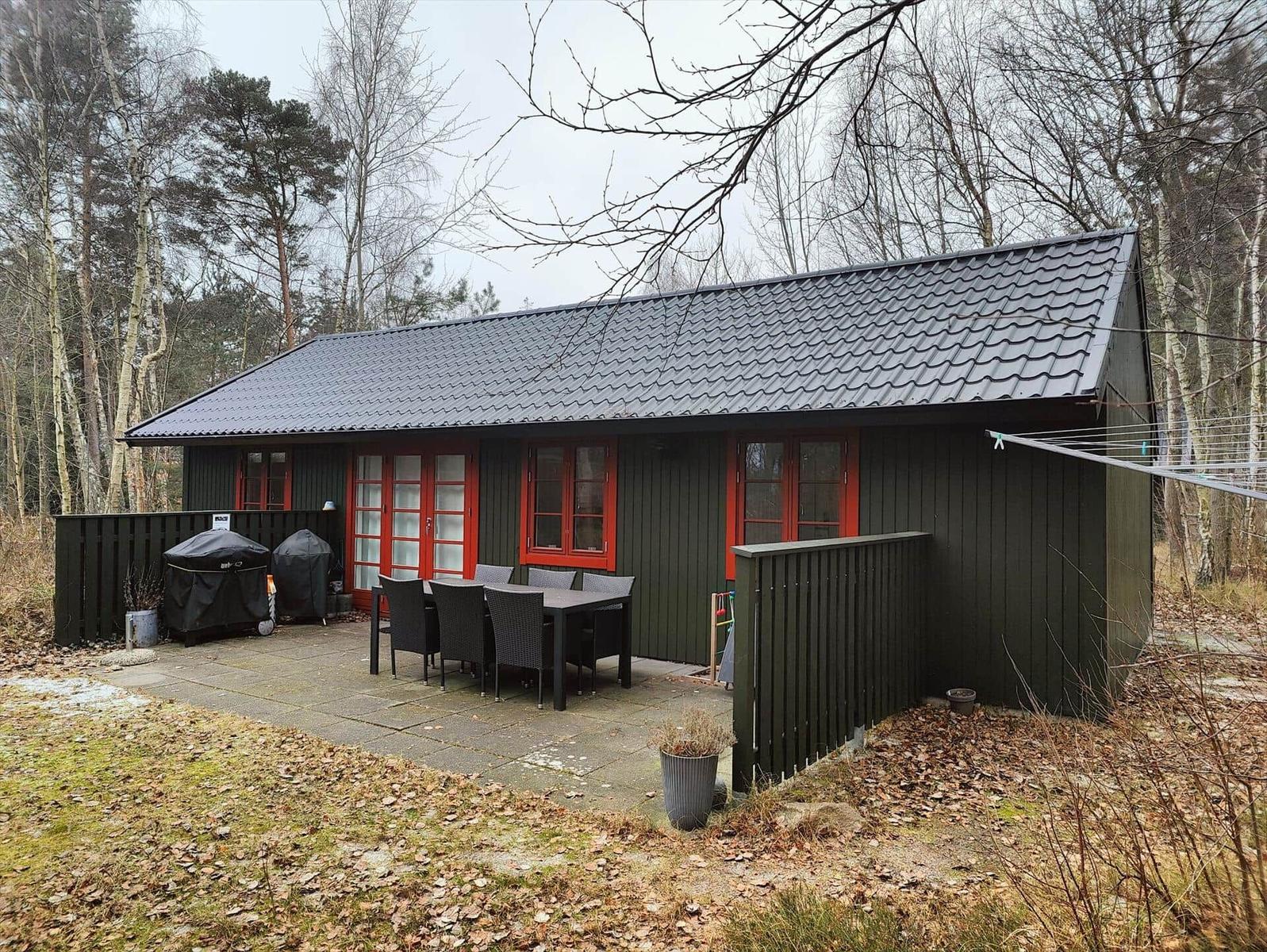 A green wooden house with red windows and a terrace with table and chairs.