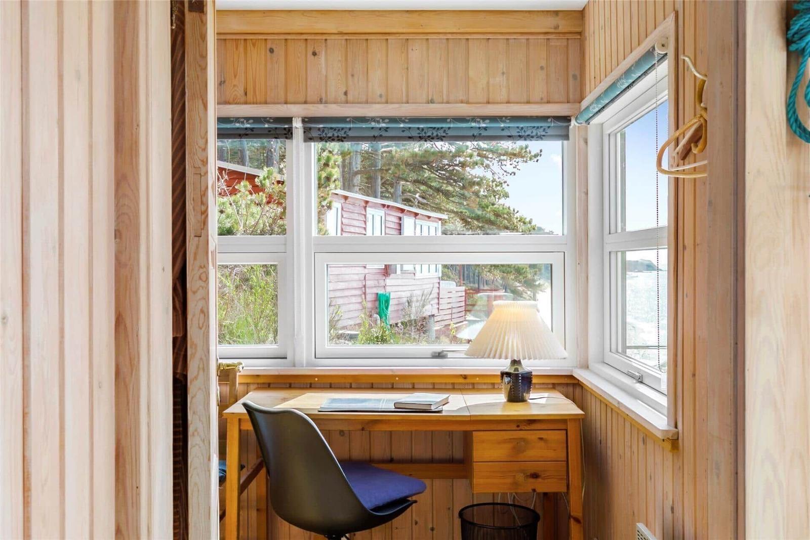 Desk with chair by window, view of wooden houses and trees.