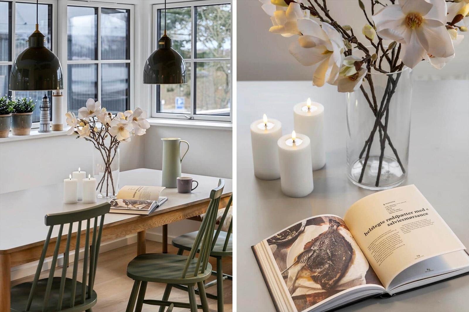 Esstisch mit Kerzen, Blumen und Buch in hellem Raum mit großen Fenstern.
