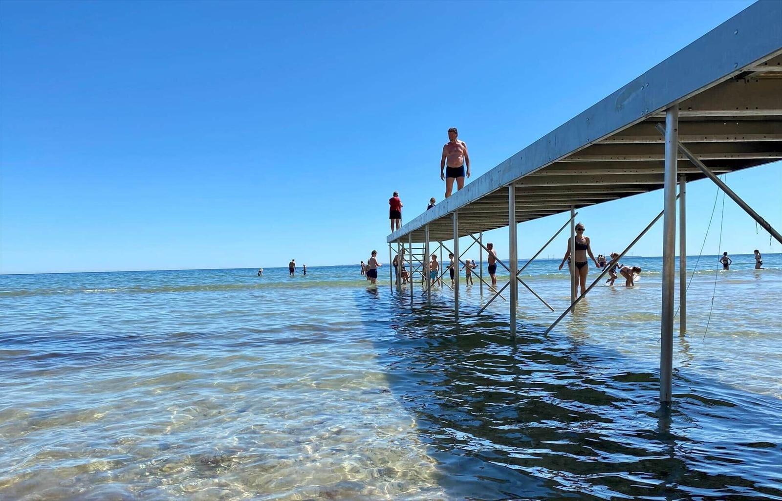 A pier extends into clear water, people enjoy swimming and sunbathing.