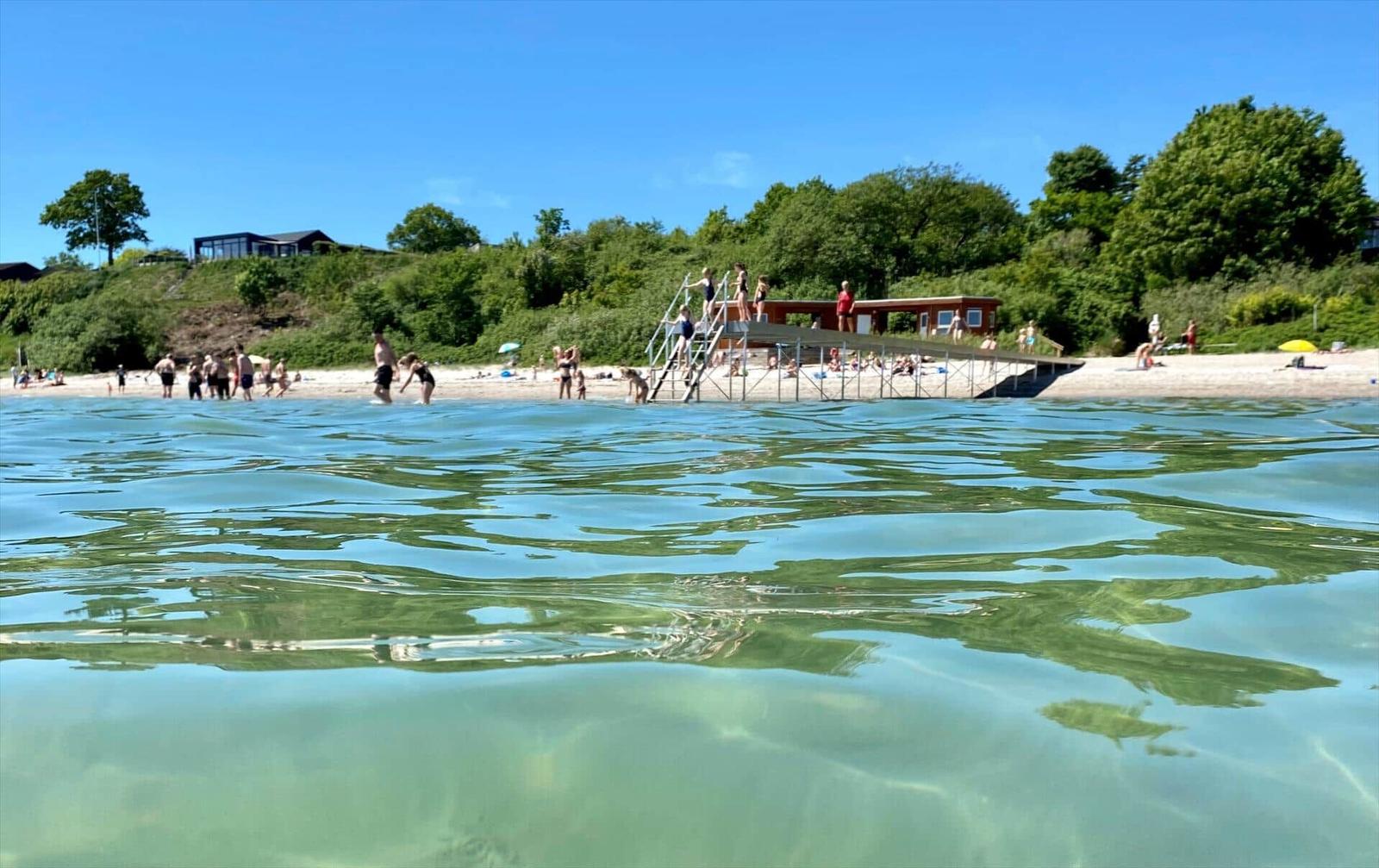 Clear water beach with pier and swimmers under blue sky.
