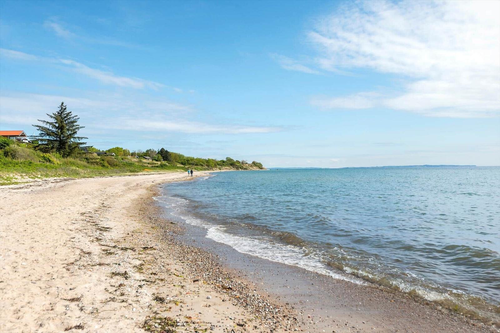 Beach with sand and stones, waves gently break. Trees and houses are visible in the background.