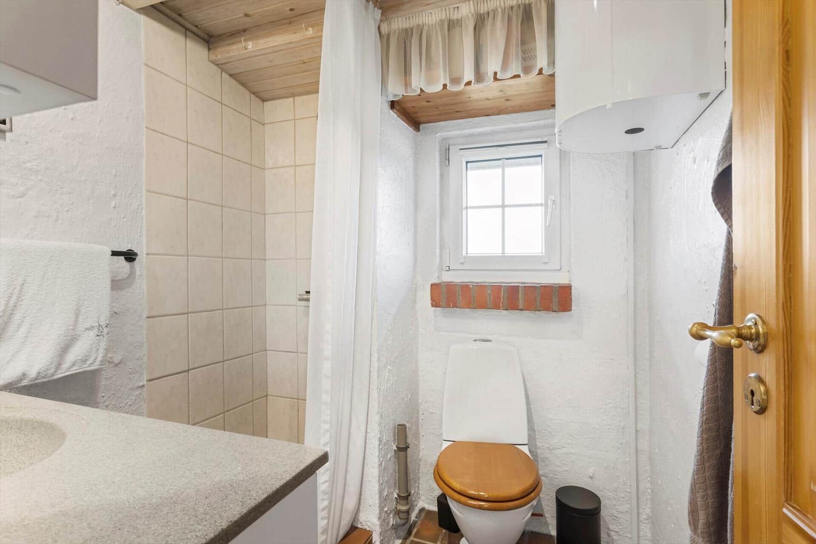 Bathroom with toilet, shower, and window. Wooden ceiling and white walls.