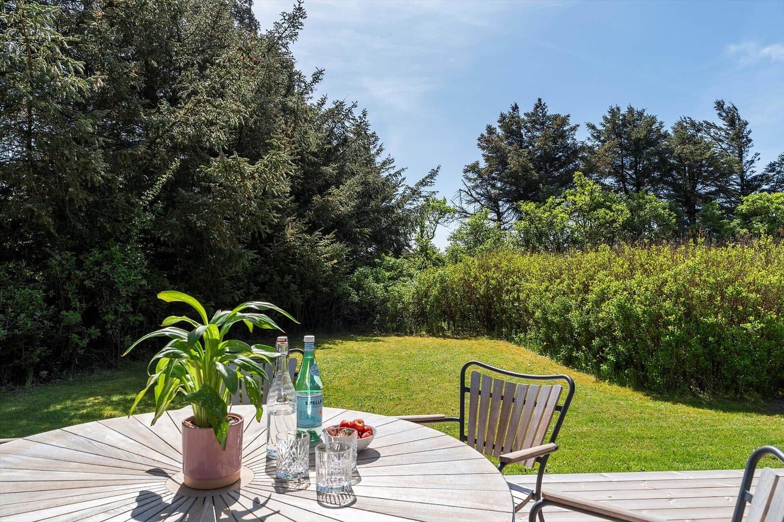 Round table with plant, glasses, and bottles on terrace with grass and trees.