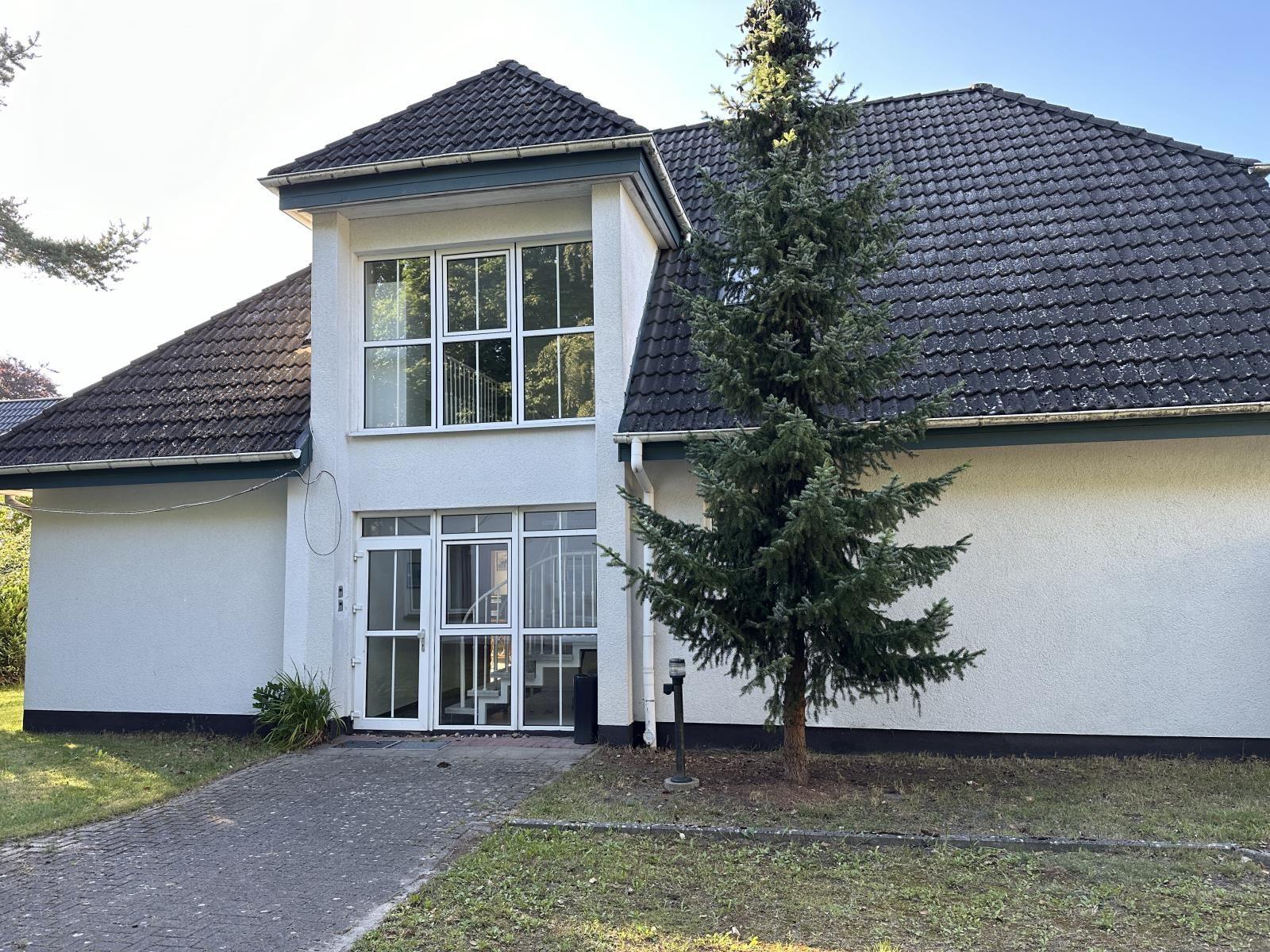 Haus mit weißer Fassade, dunklem Dach und Glasfront. Vorgarten mit Baum und Kiesweg.