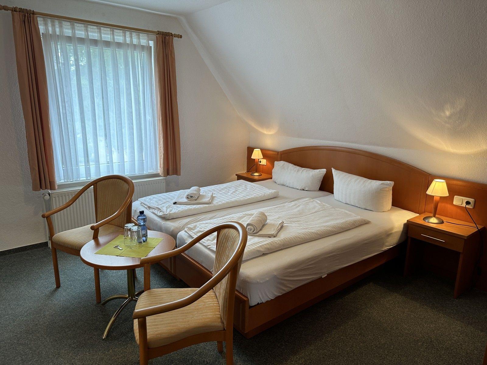 Room with double bed, two nightstands, chair and table. Window with curtains.