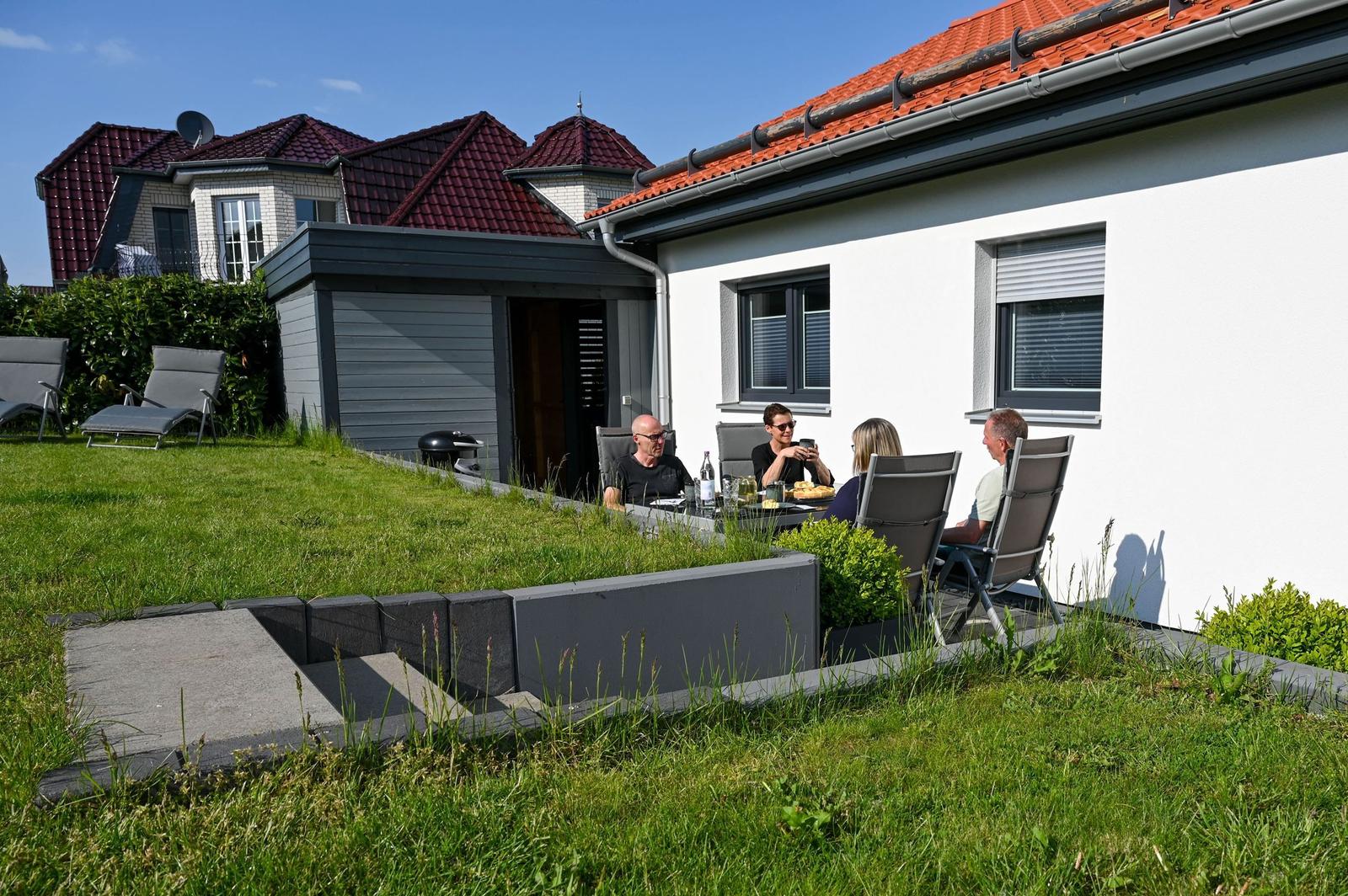 Grüner Garten mit Terrasse und Grill. Gäste sitzen an Tisch. Haus mit rotem Dach.