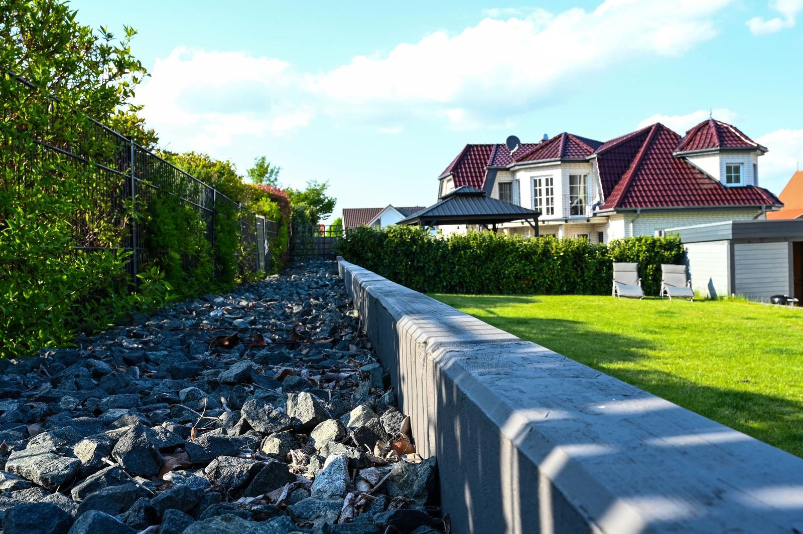 Garten mit Rasenfläche, Steinschüttung und Steinmauer. Im Hintergrund Haus mit rotem Dach.
