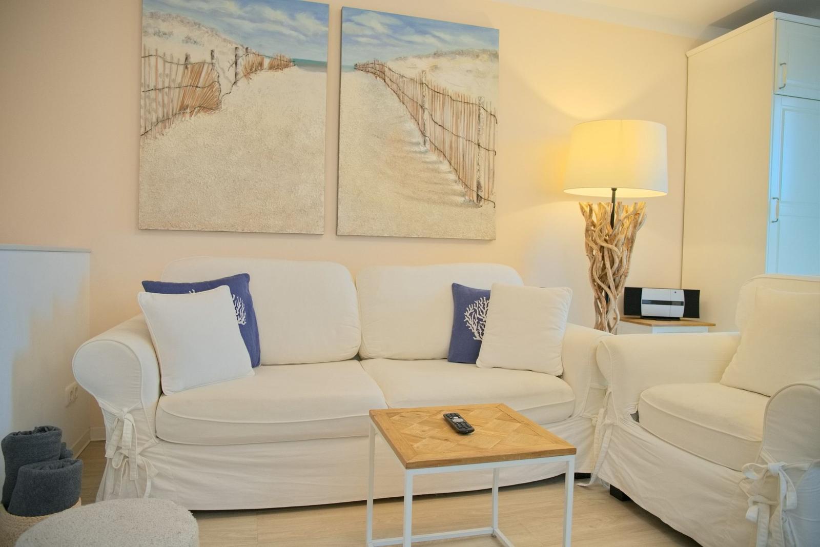 Wohnzimmer mit Sofa, Sessel und Tisch. Wandbilder zeigen Strand. Stehlampe mit Holzgestell.