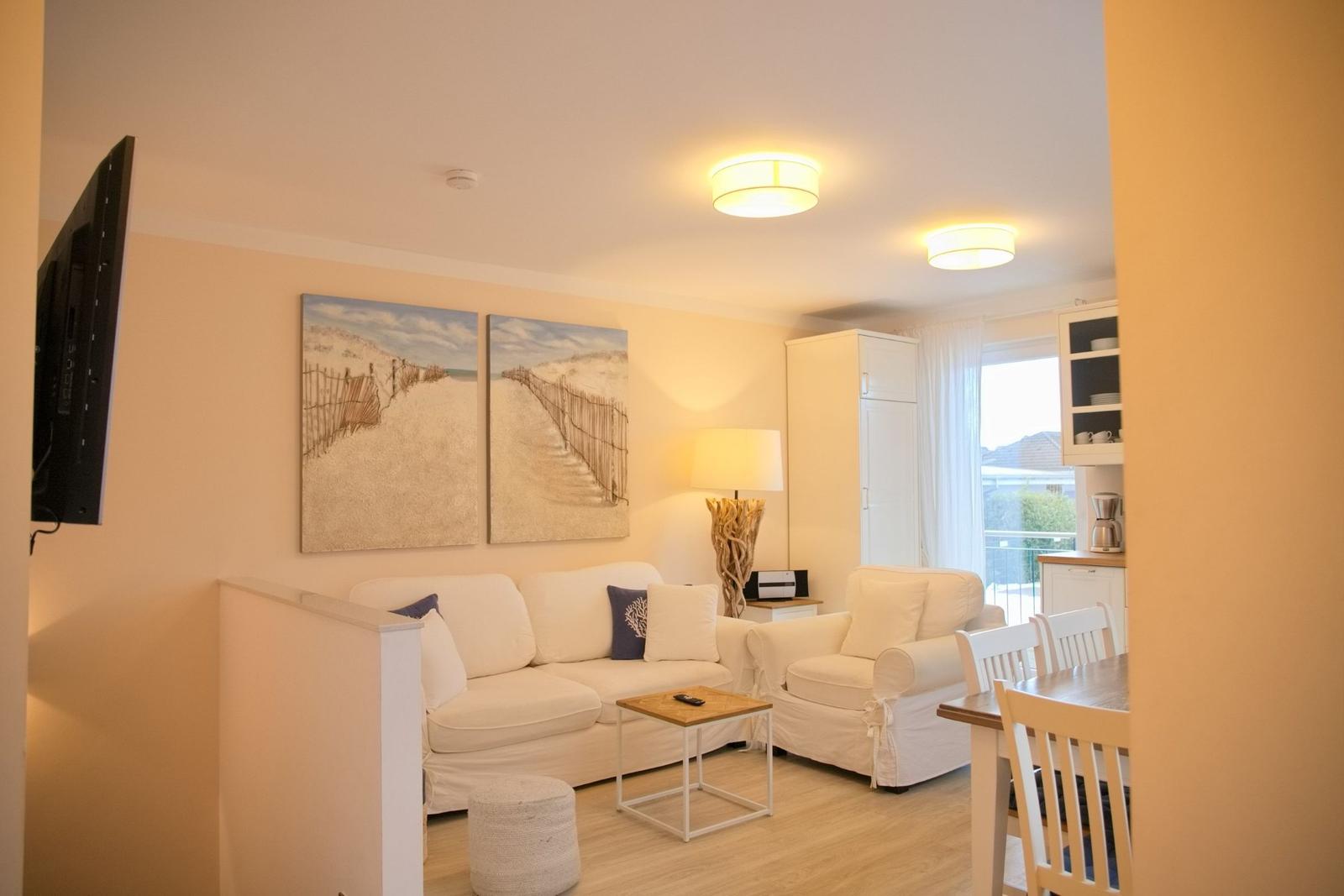 Wohnzimmer mit Sofa, Sessel und TV. Wandbilder zeigen Strand. Küchenbereich im Hintergrund.