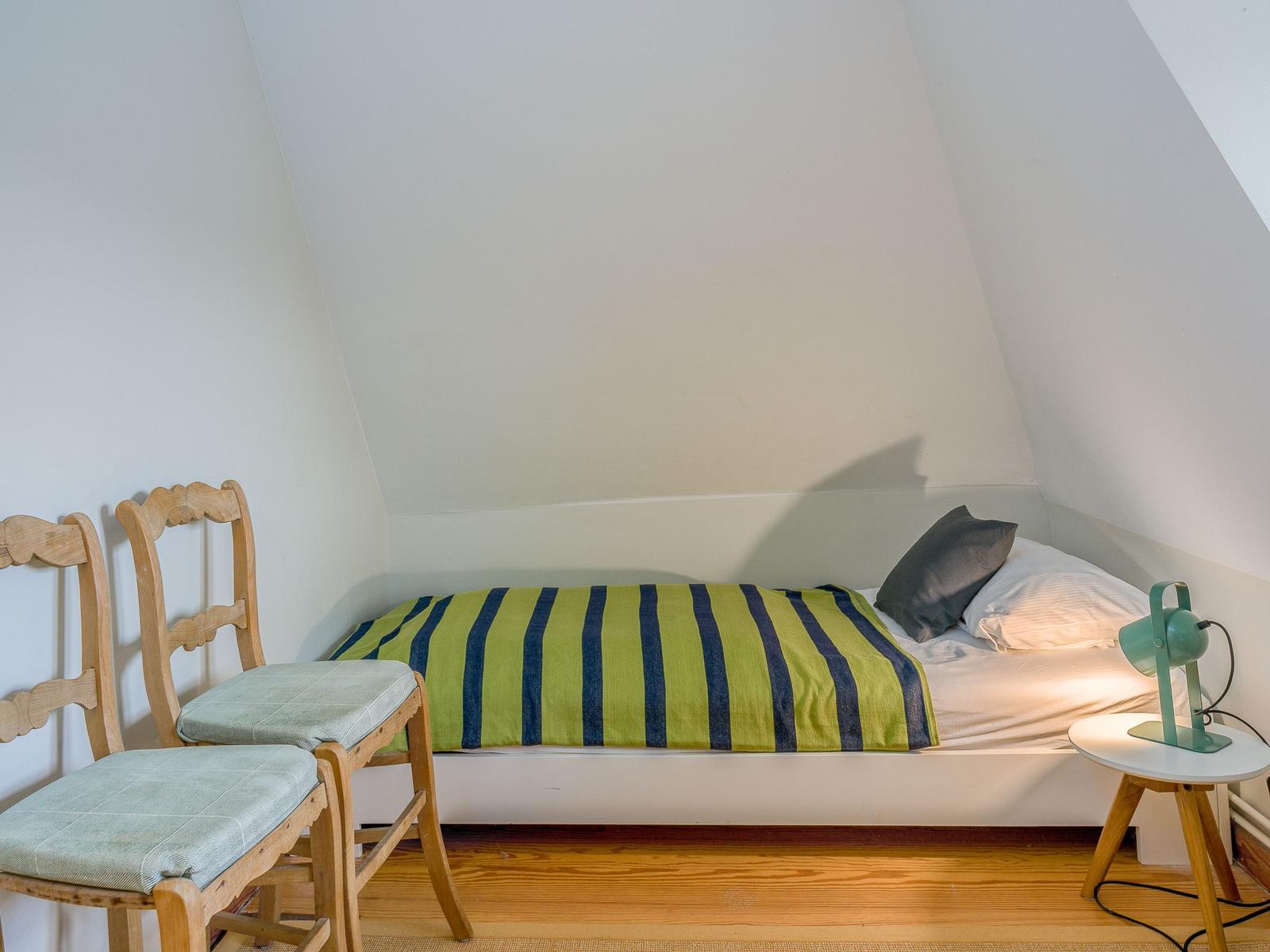 Ein Schlafzimmer mit Dachschrägen, Bett, zwei Stühlen und Nachttisch mit Lampe.