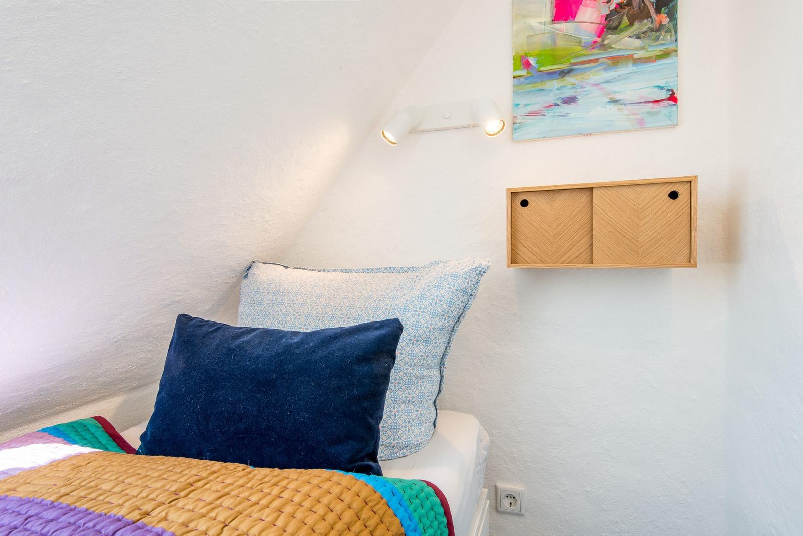 Schlafzimmer mit Bett, Kissen und Wandregal. An der Wand hängt ein Bild und eine Lampe.