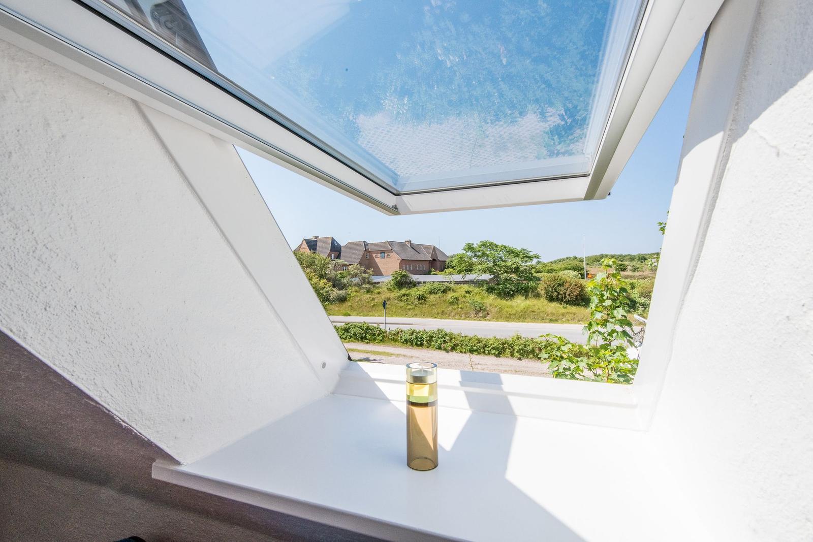 Dachfenster mit Blick auf Straße und Häuser. Auf Fensterbank steht eine Glasflasche.