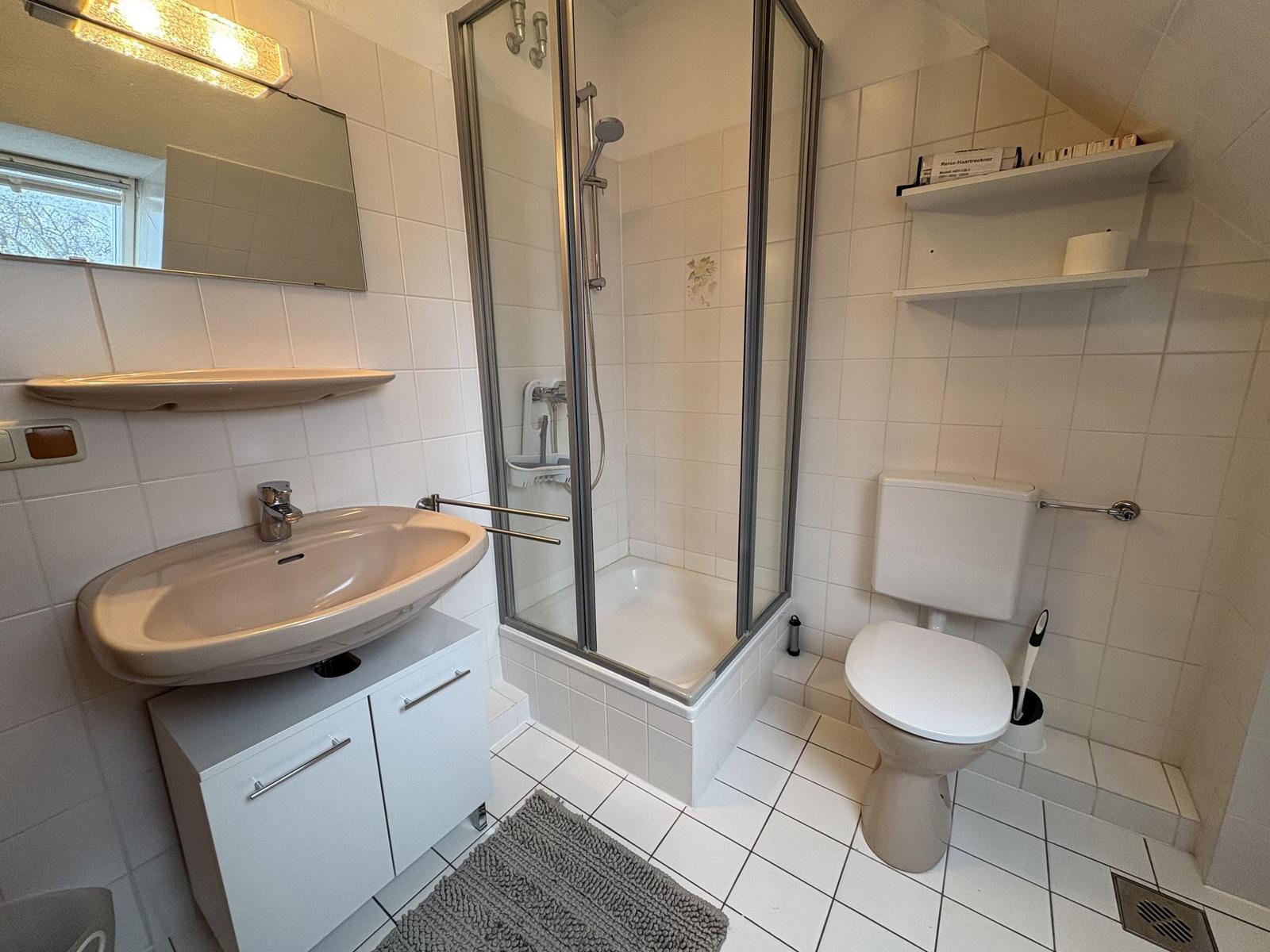 Badezimmer mit Dusche, Waschbecken und Toilette. Weiße Fliesen und Spiegel.