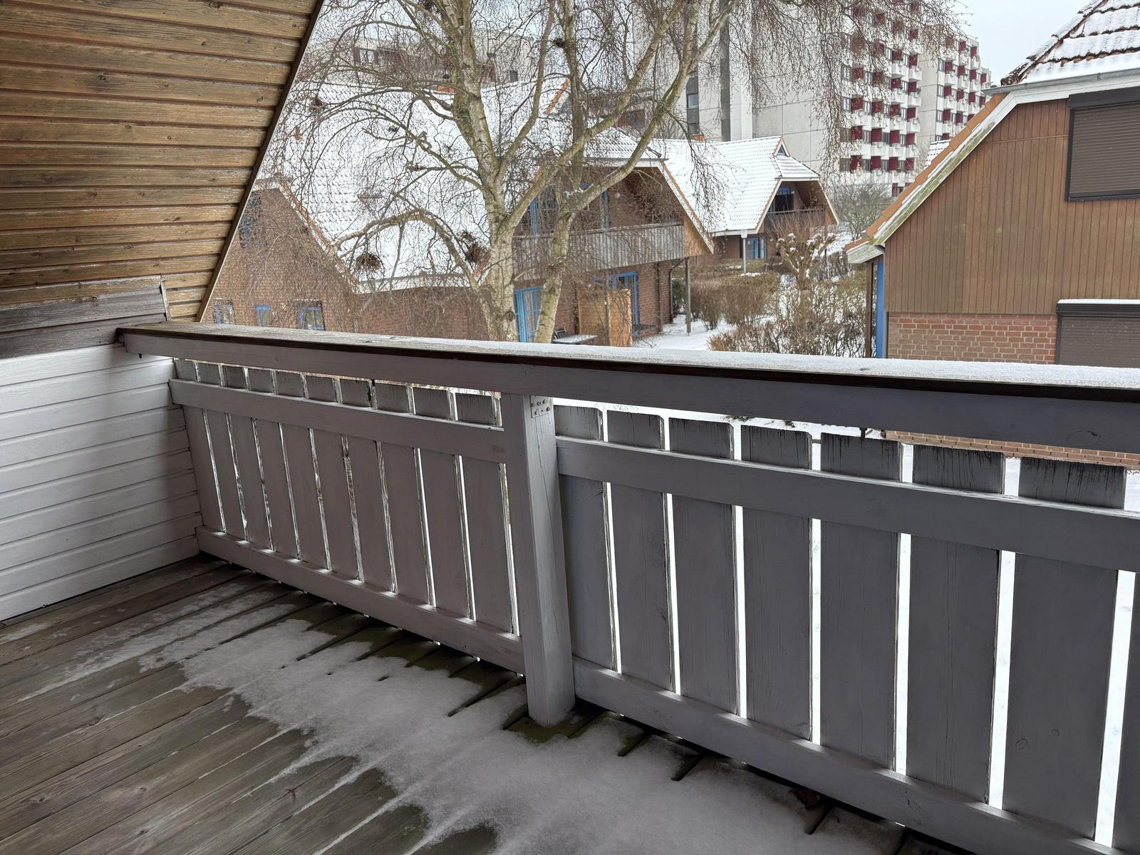 Balkon mit Holzgeländer und Schnee, Blick auf verschneite Häuser und Bäume.