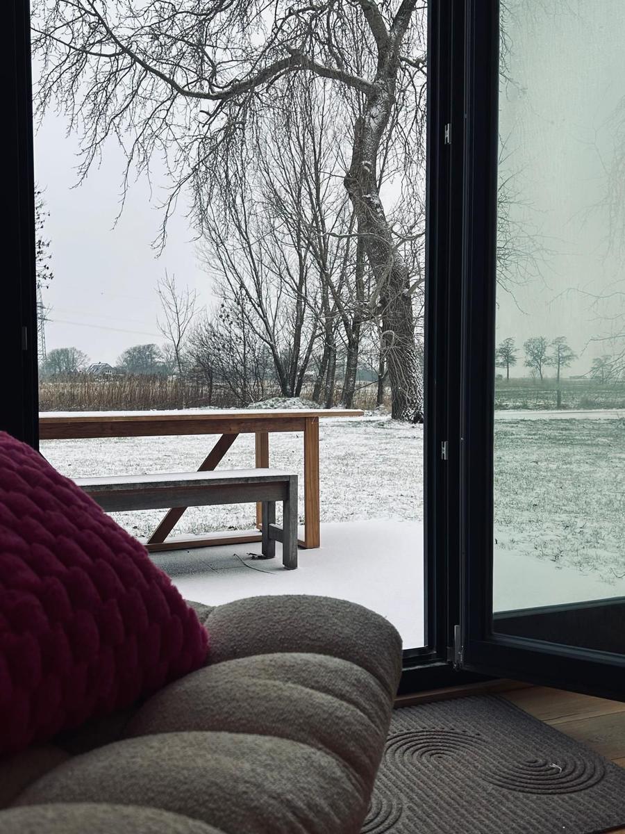 Durch große Fenster blickt man auf eine schneebedeckte Terrasse mit Holztisch und Bank.