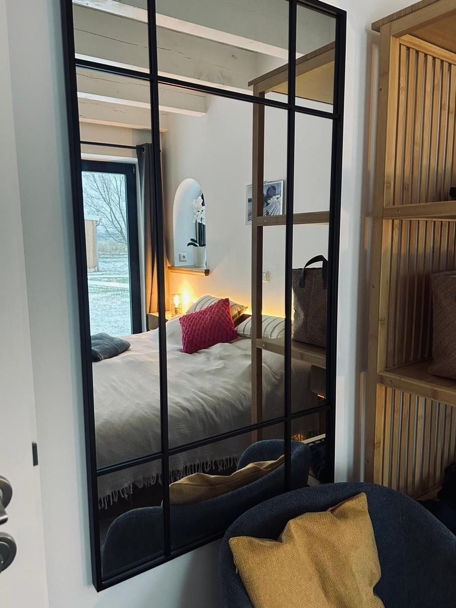 Schlafzimmer mit Bett, Spiegel und Holzregal. Fenster zeigt Außenbereich.