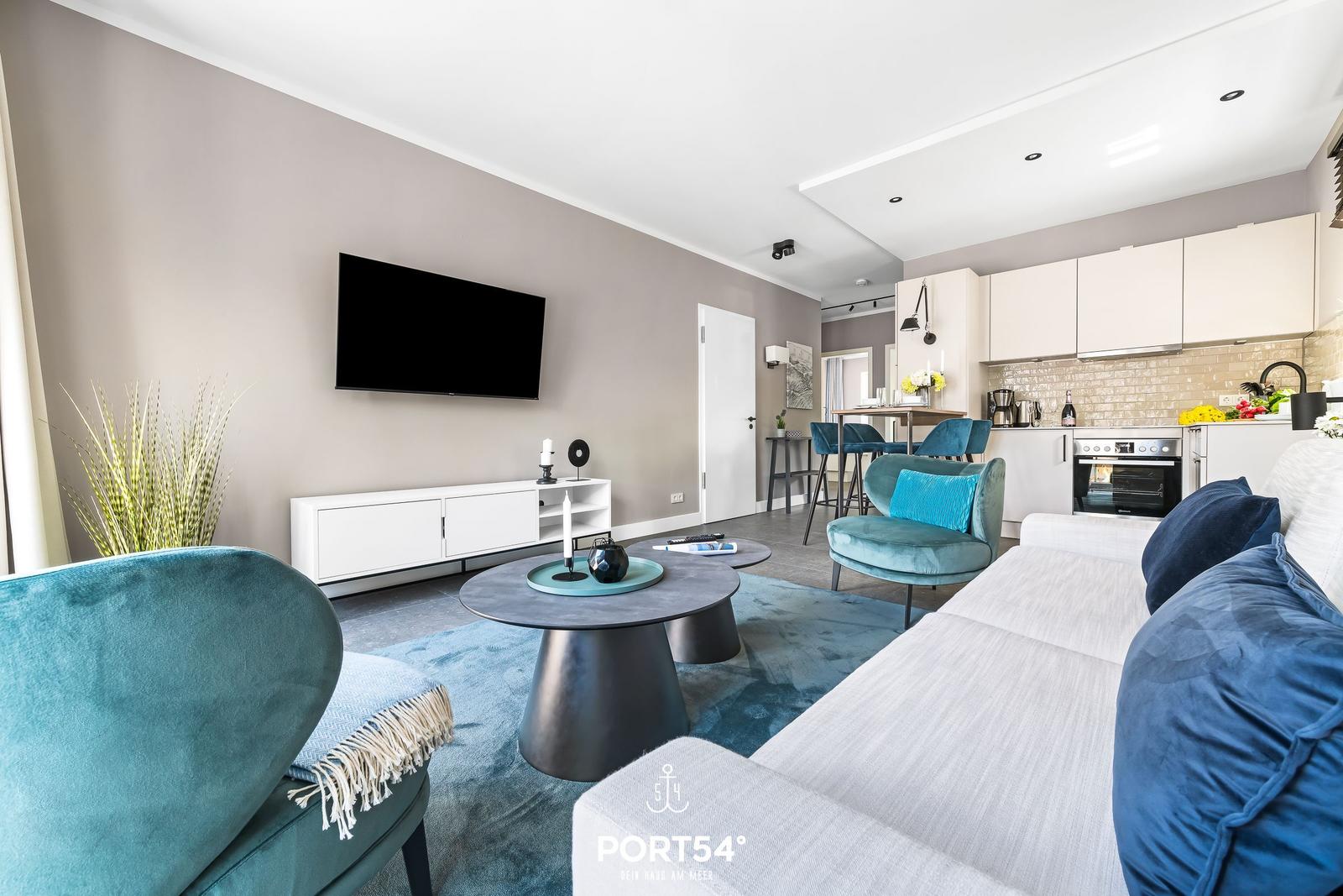 Wohnzimmer mit Sofa, Sessel und Fernseher. Küche mit Arbeitsplatte und Kochgeräten.