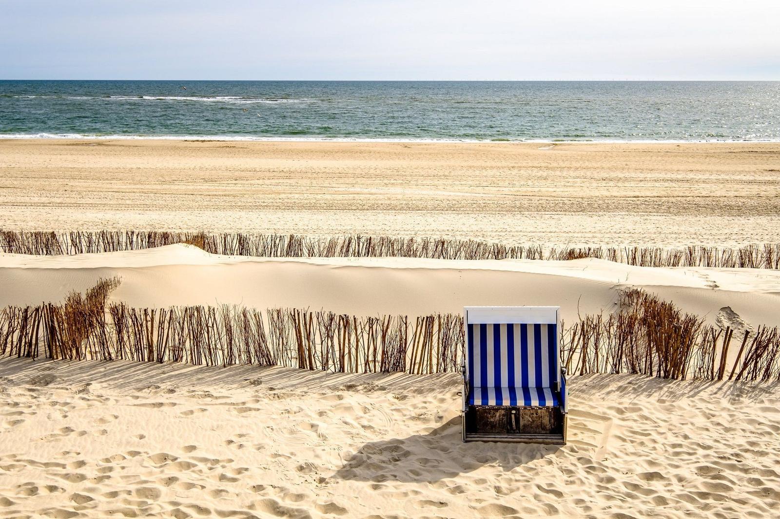 Ein blaues Strandkörbchen steht auf sandigen Dünen vor dem Meer.