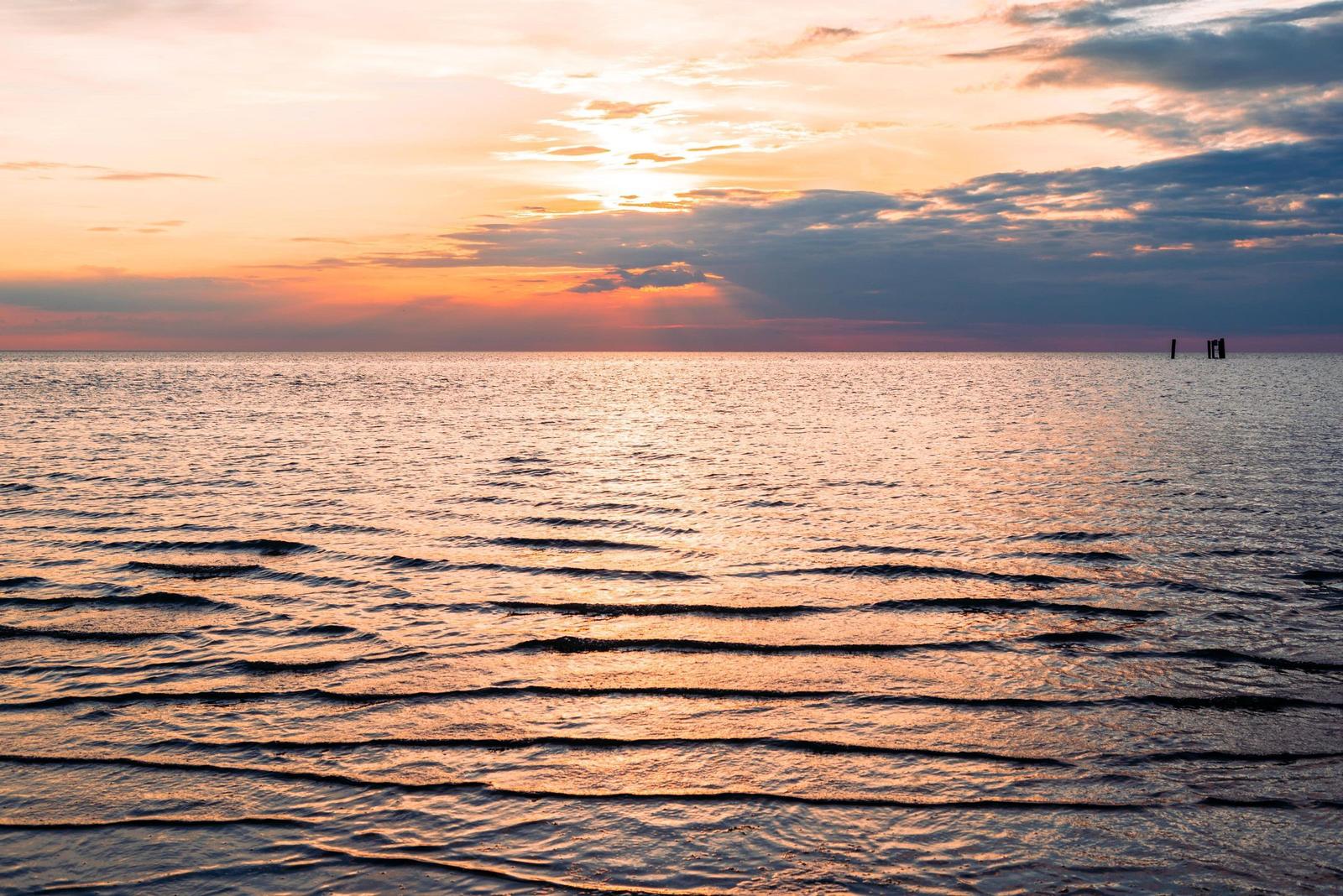 Sonnenuntergang über ruhigem Wasser mit sanften Wellen und Wolken am Horizont.